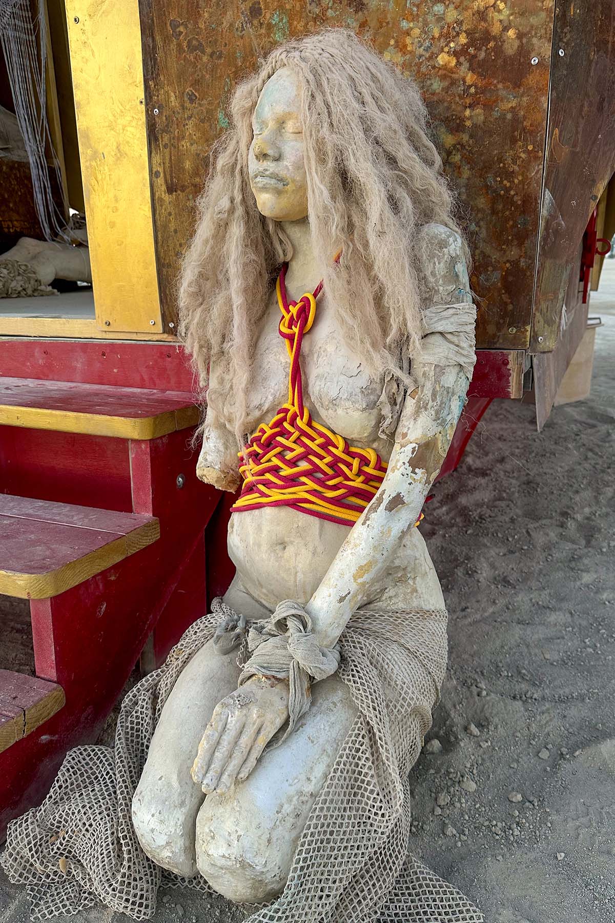 Sculpture of a kneeling woman with long pale hair and a red woven top, seated beside the red steps of the pavilion