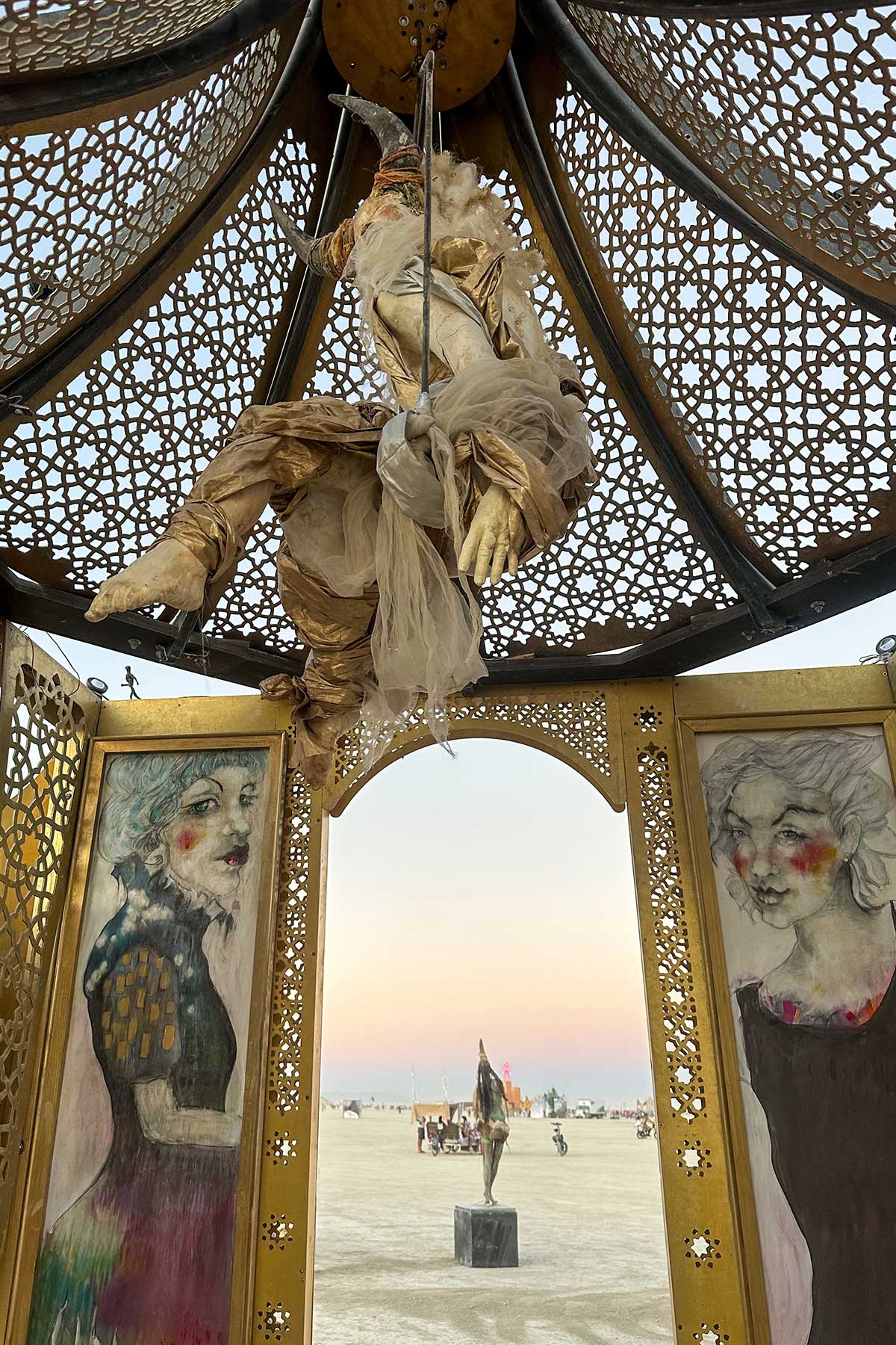 Looking up inside the pavilion at a draped figure suspended beneath the ornate lattice dome, with painted portraits framing the arched doorway