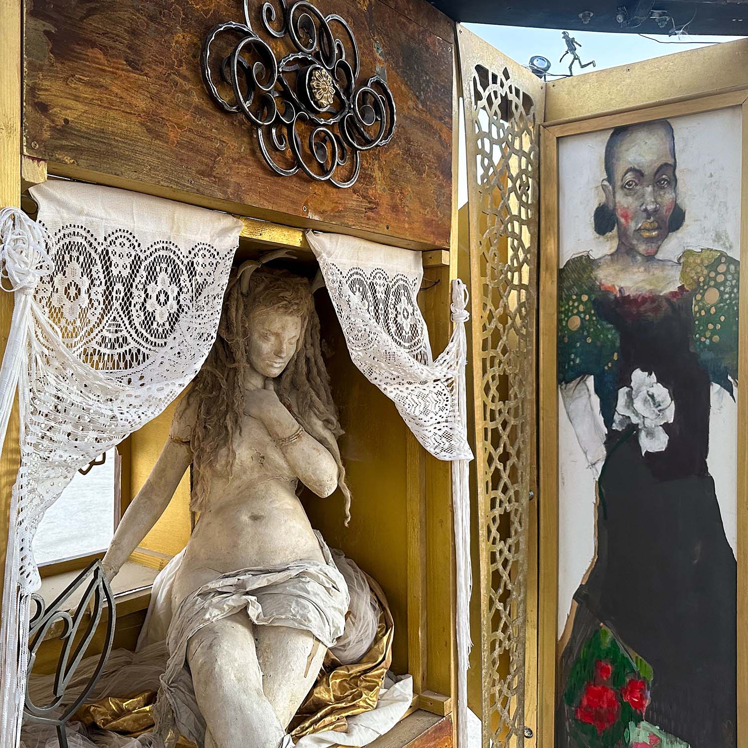 Sculpted reclining woman in an alcove framed by lace curtains, beside a painted portrait of a woman in a dark dress