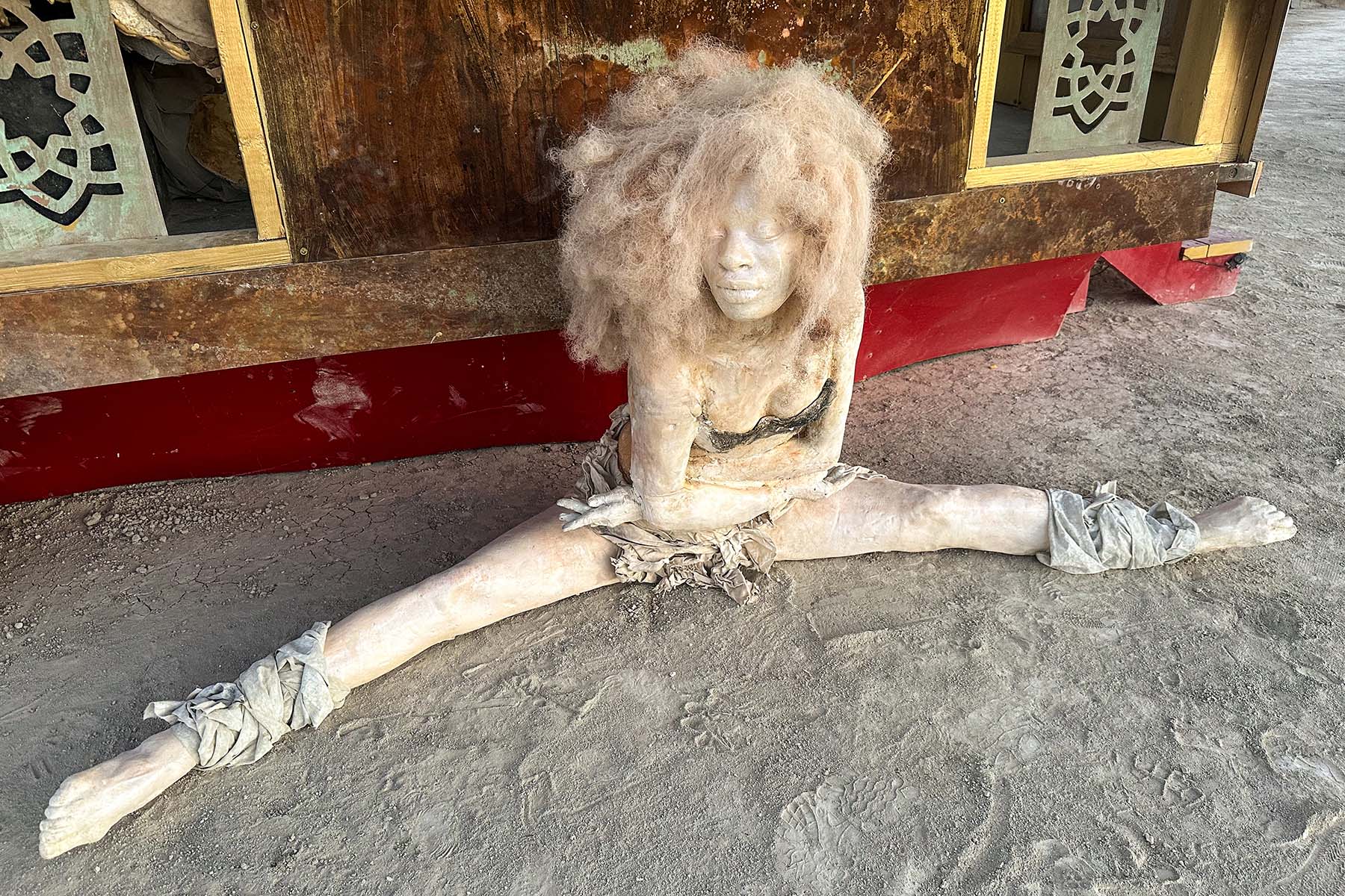 Sculpture of a woman with wild pale hair doing the splits on the playa in front of the pavilion
