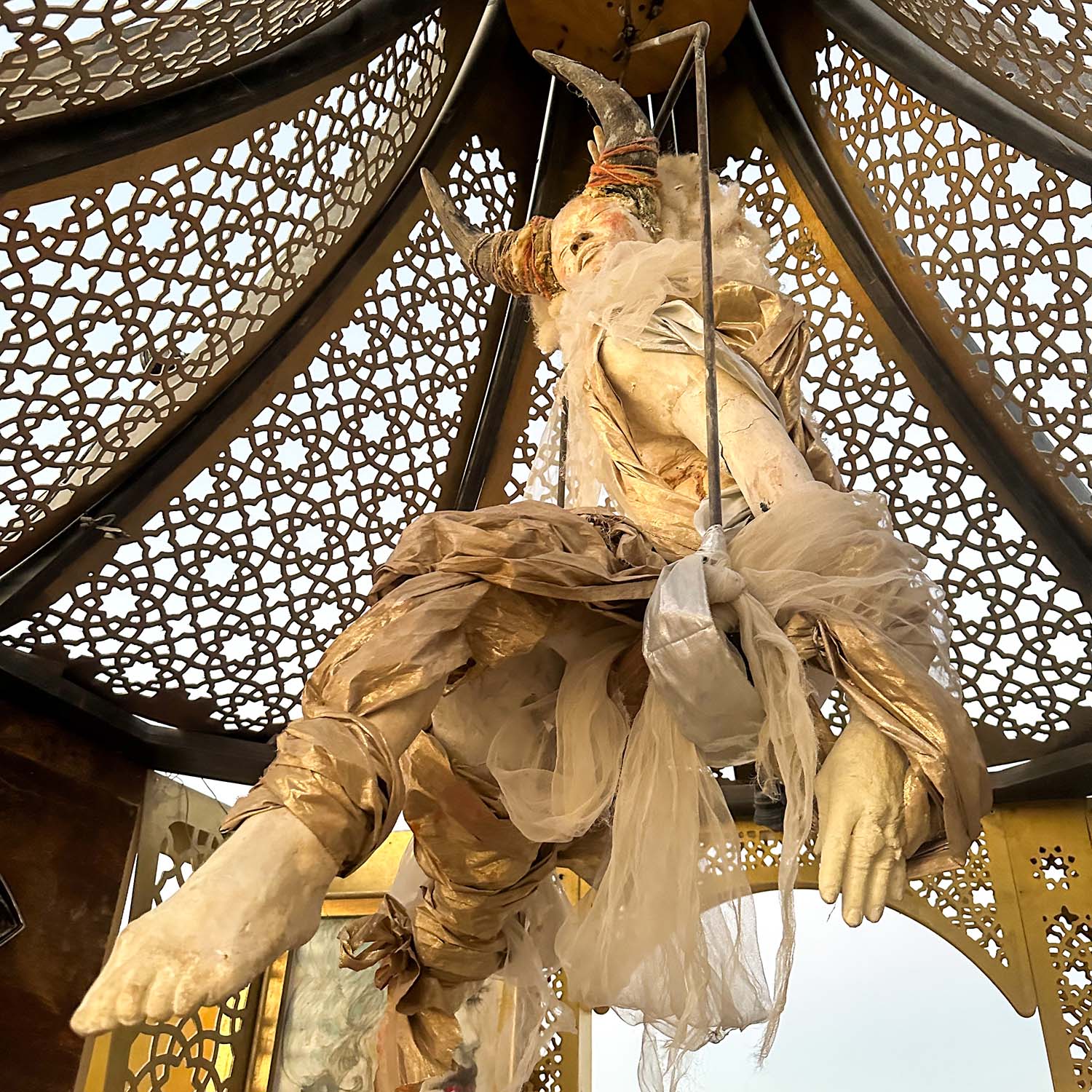 A cloth-draped figure suspended from the lattice dome inside the pavilion, seen from below
