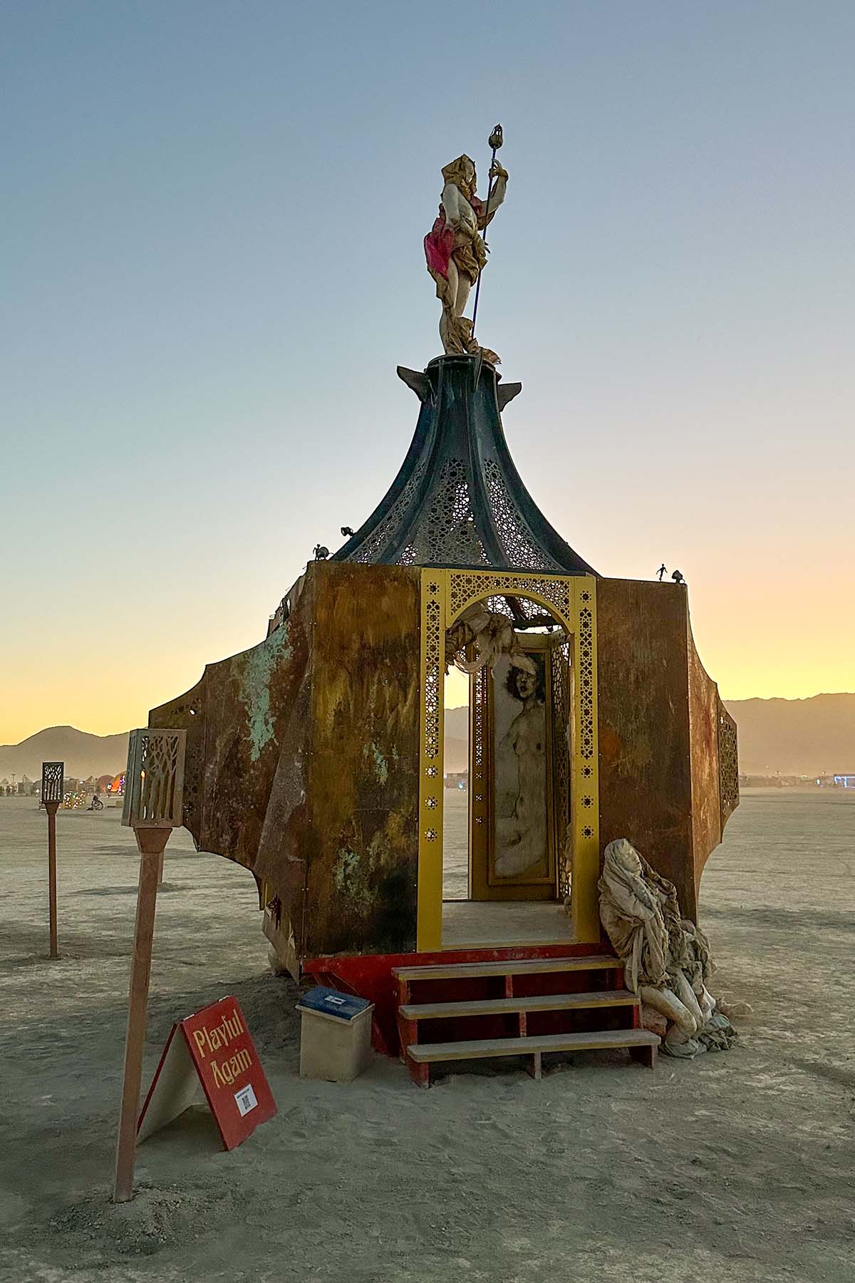 The pavilion at sunset, its red steps leading to a gold-framed doorway, with a kneeling sculpture beside it