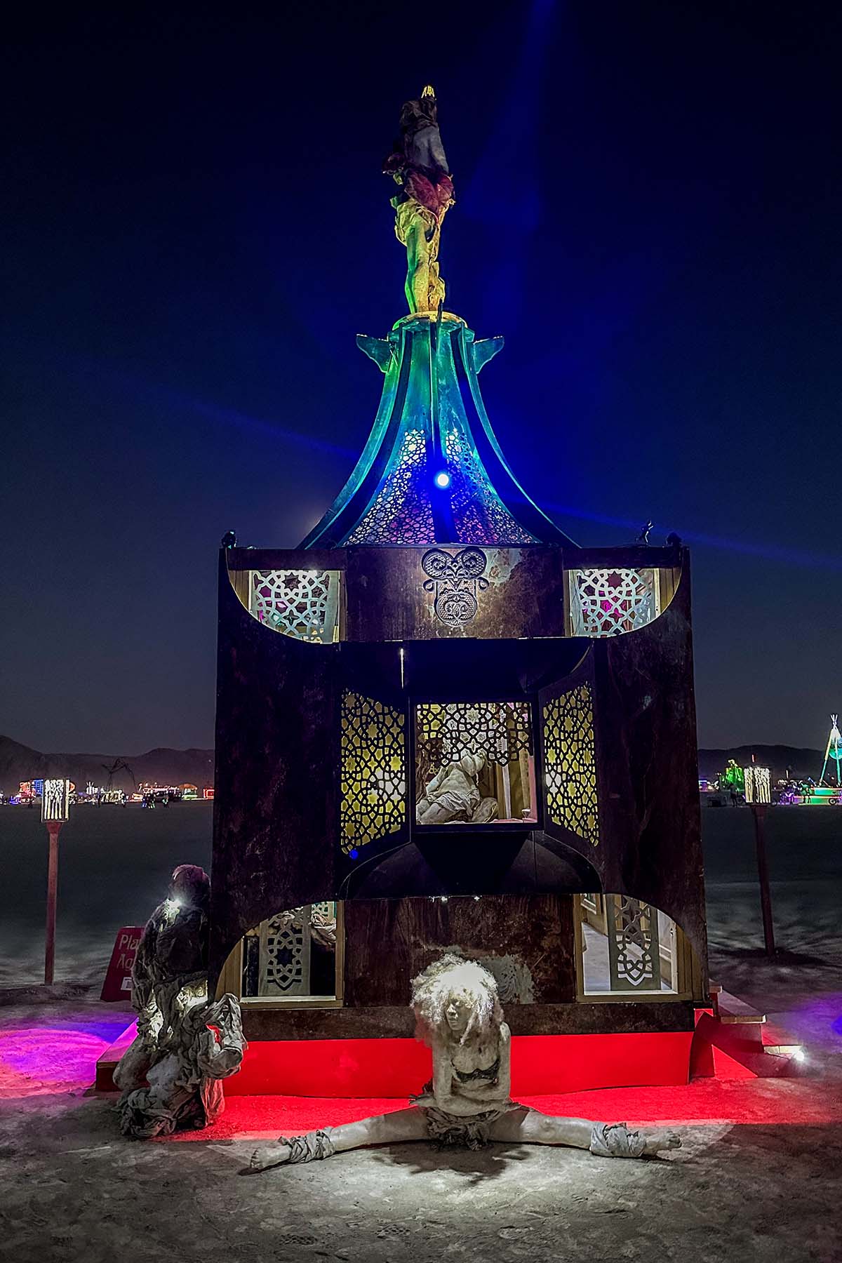 Night view of the pavilion's back, glowing green and blue above a red-lit base with figures at its feet