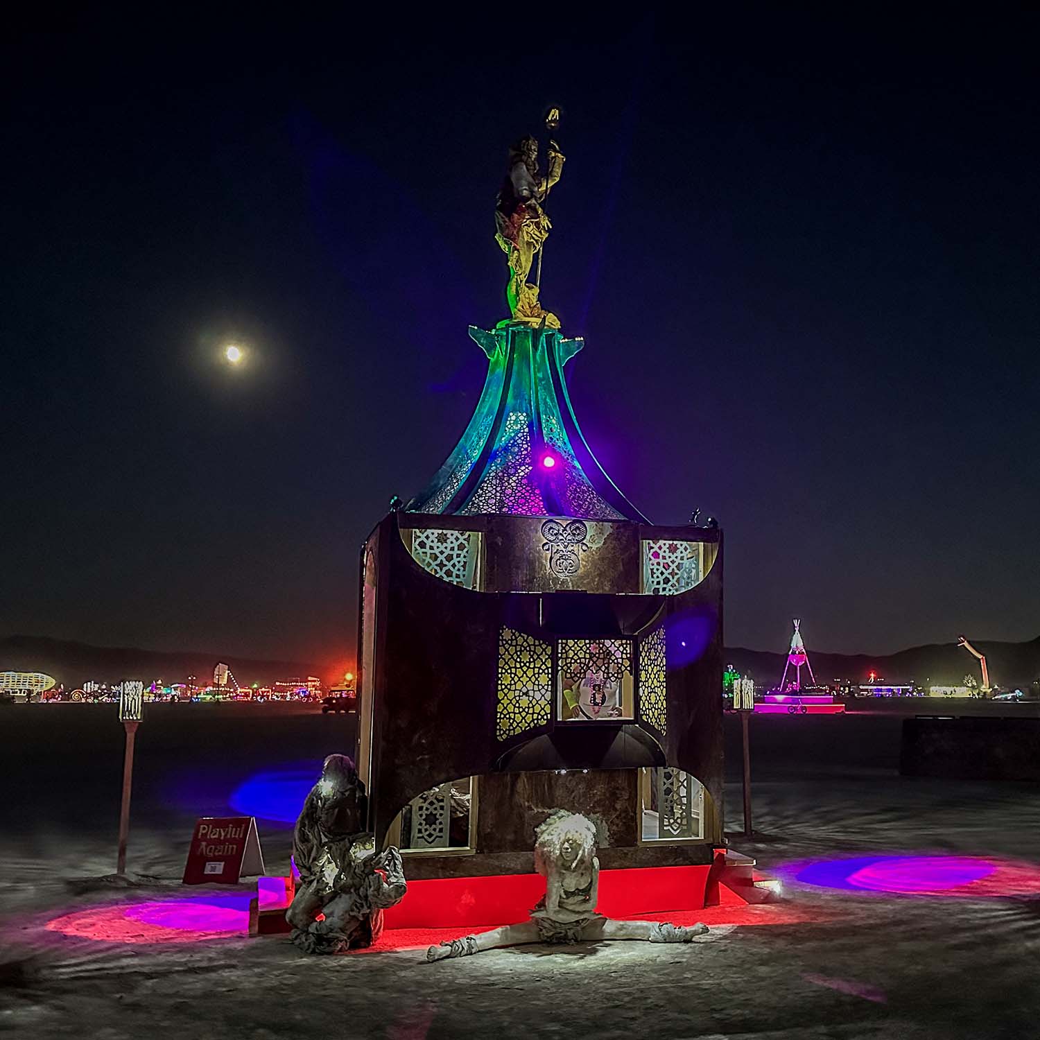 Close night view of the illuminated pavilion under the moon, light pooling red and purple on the playa
