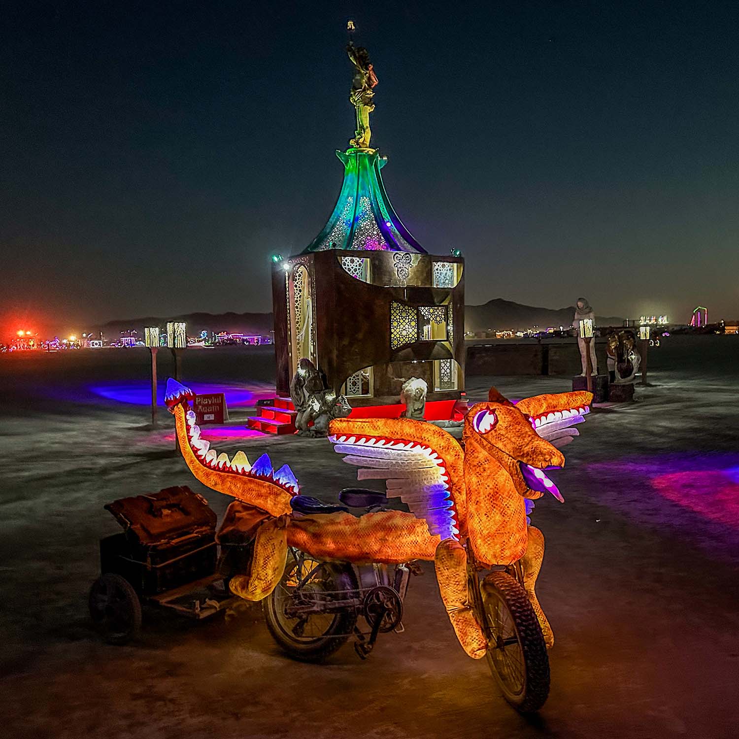 A glowing dragon-shaped art car parked in front of the illuminated pavilion at night