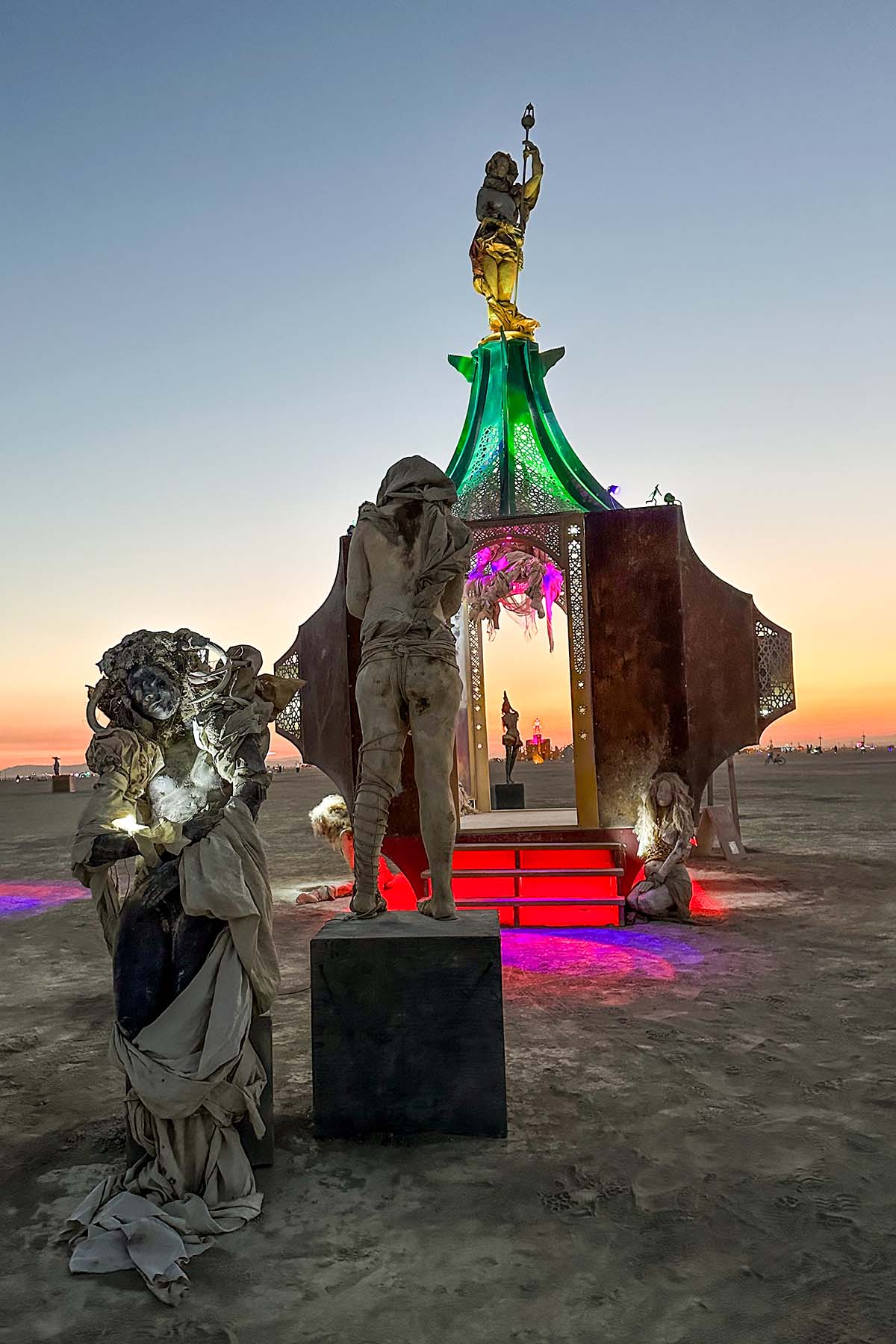 Hooded sculptures gathered in front of the glowing pavilion at sunset