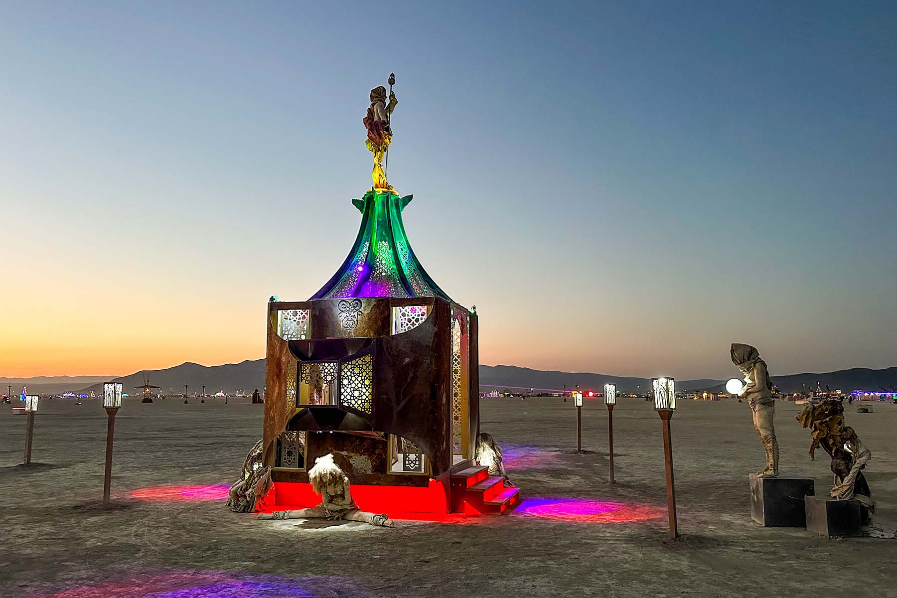 Playful Again at dusk, surrounded by sculptures and lantern posts with mountains in the distance