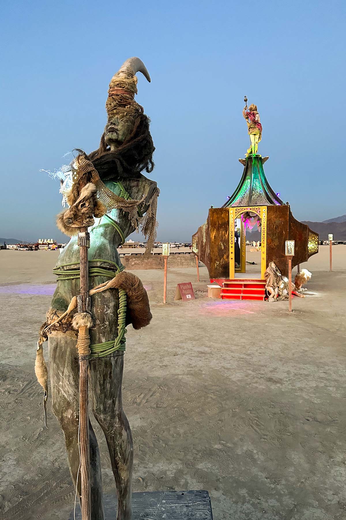 The horned figure with its staff seen from behind, looking toward the lit pavilion
