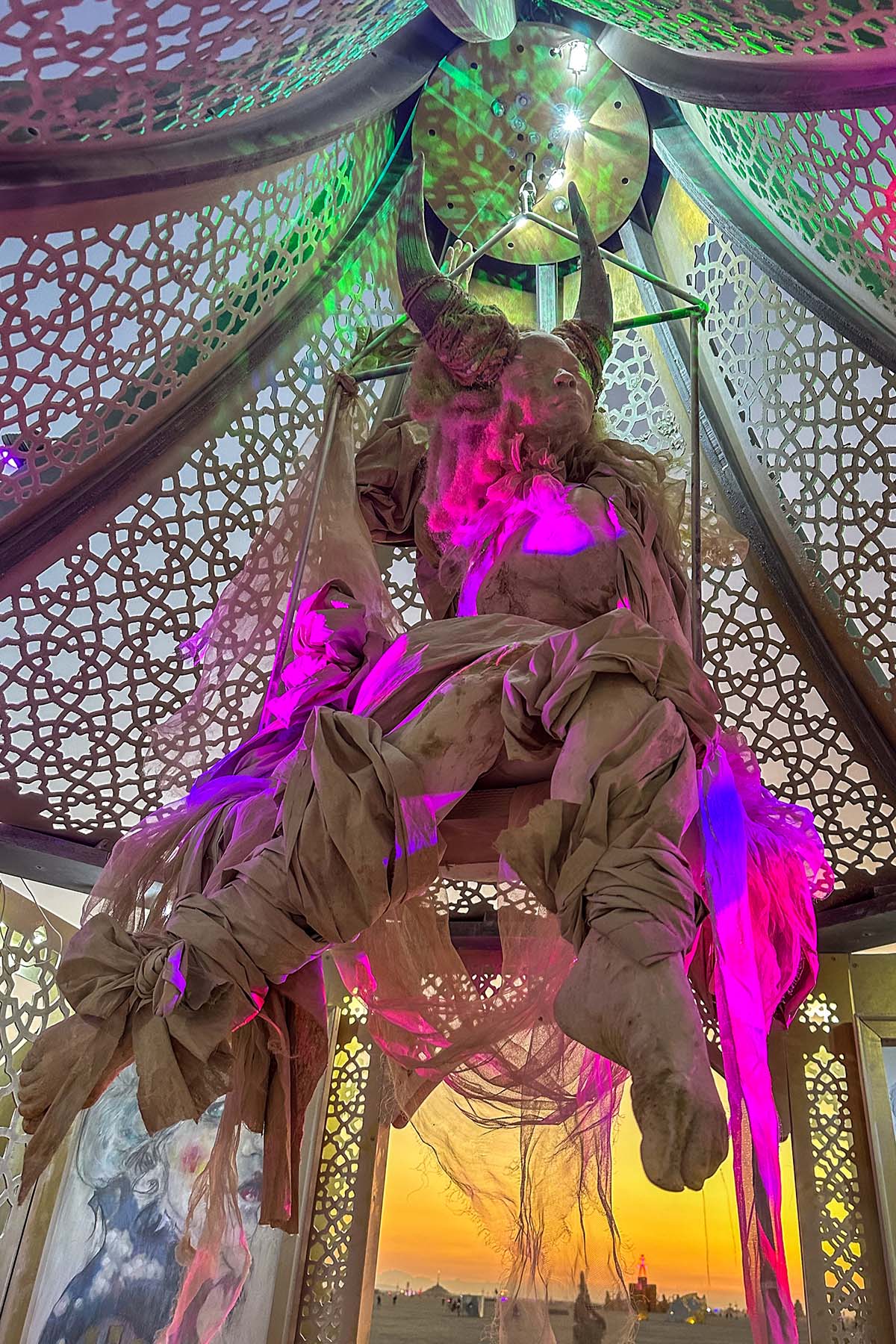 A draped figure suspended under the dome, lit pink and purple, with the sunset glowing through the arch below