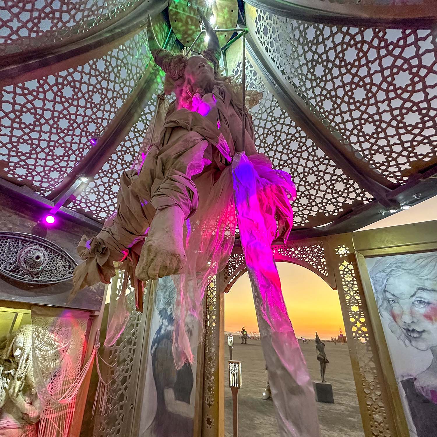 Inside the pavilion at sunset, a draped hanging figure lit in pink and purple above the doorway