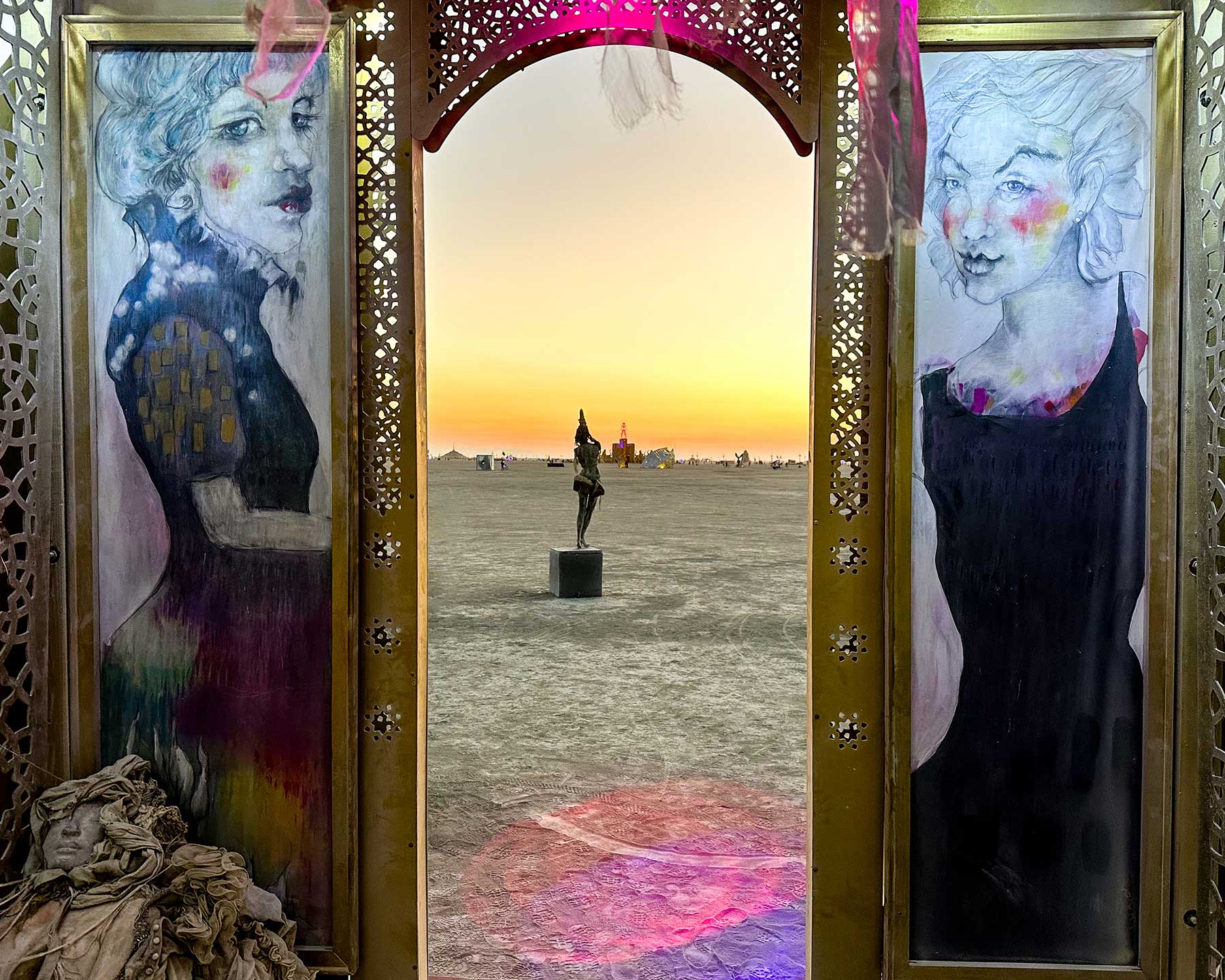 Painted portraits of two women framing the arched doorway, with a sculpture visible on the playa at sunset