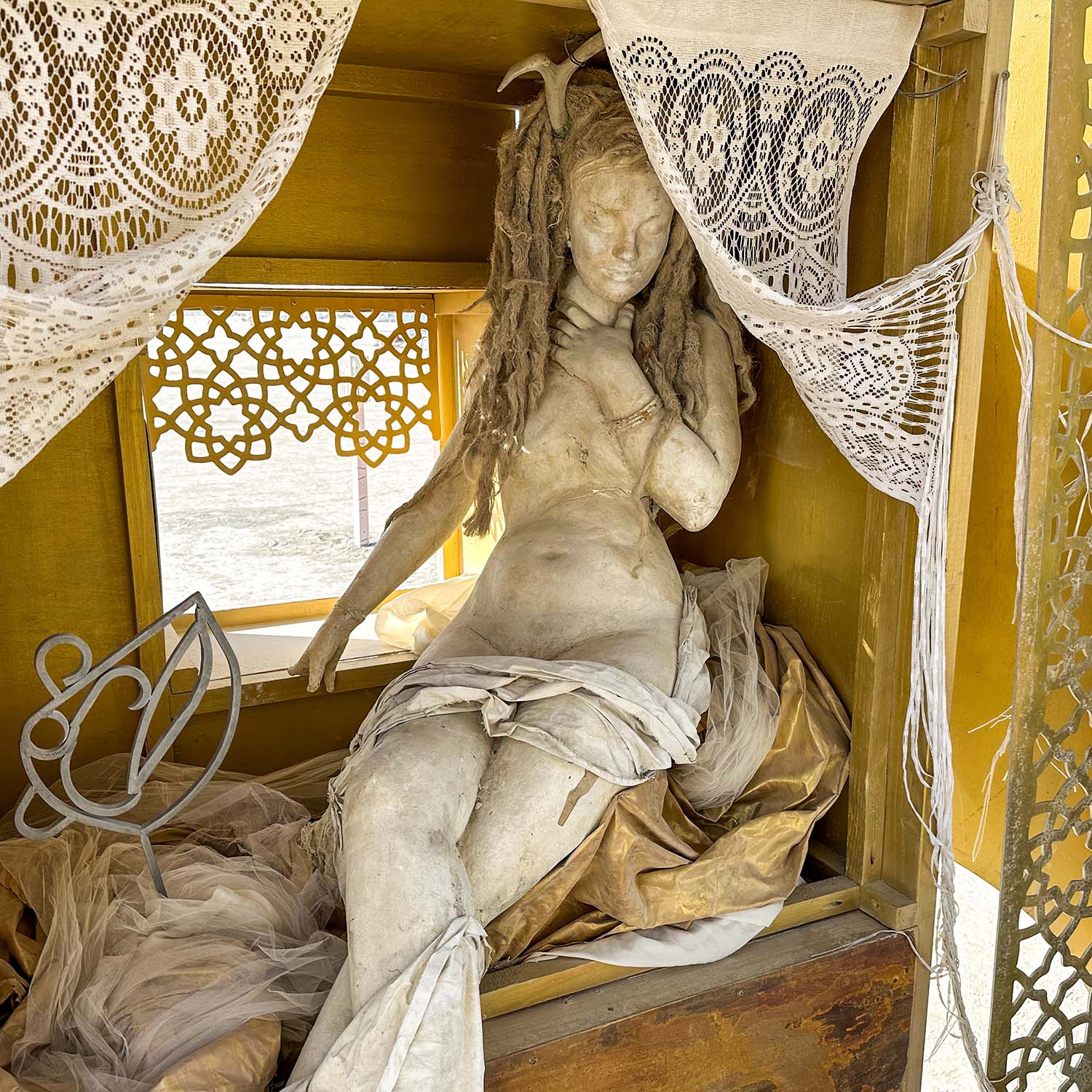 Sculpted reclining woman in a golden alcove framed by white lace curtains, in daylight