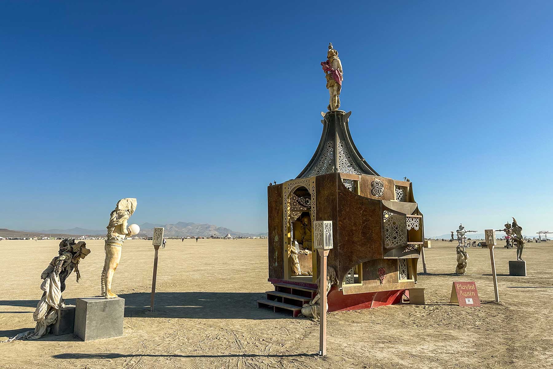 Playful Again by day: an ornate pavilion topped by a dancing figure, with cast figures and lamp posts around it on the playa